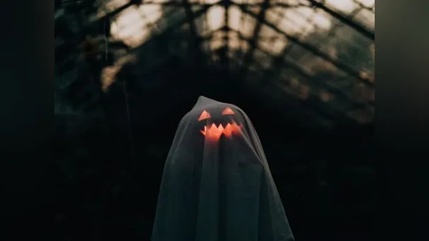 A glowing jack-o’-lantern face shines through a white ghost costume in a dark setting.