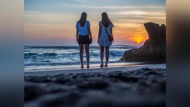 Two women stand at the shoreline, watching the sun set over the ocean.