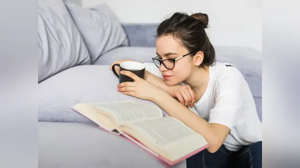 Woman with glasses holds a mug while reading an open book beside a sofa.